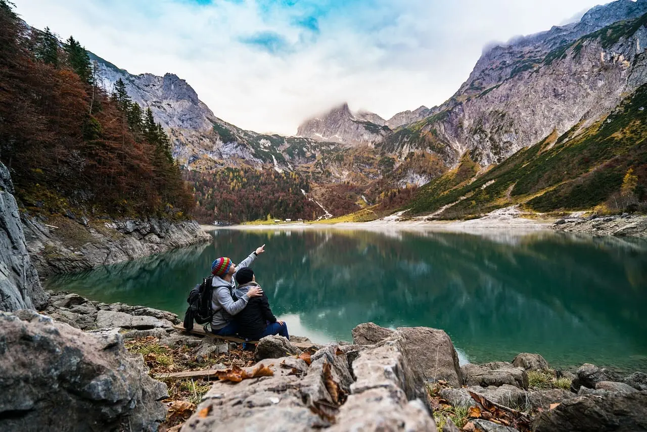 A couple sitting on rocky terrain near a calm mountain lake, surrounded by towering peaks with autumn-colored trees. One person is pointing towards the mountains, while the other leans in closely, enjoying the scenic view