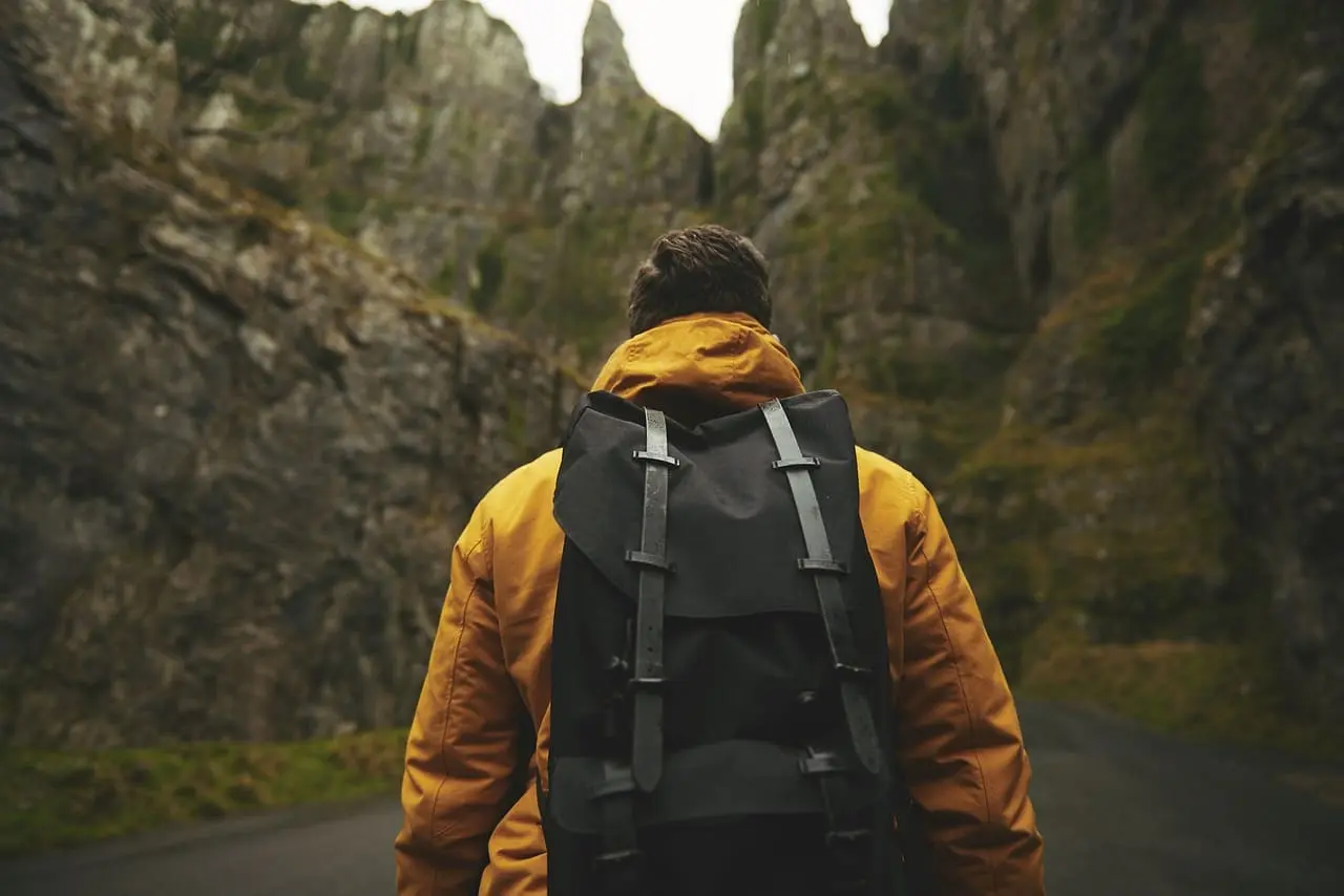 A traveler wearing a yellow jacket and black backpack walking through a narrow mountain pass, surrounded by towering cliffs and lush greenery