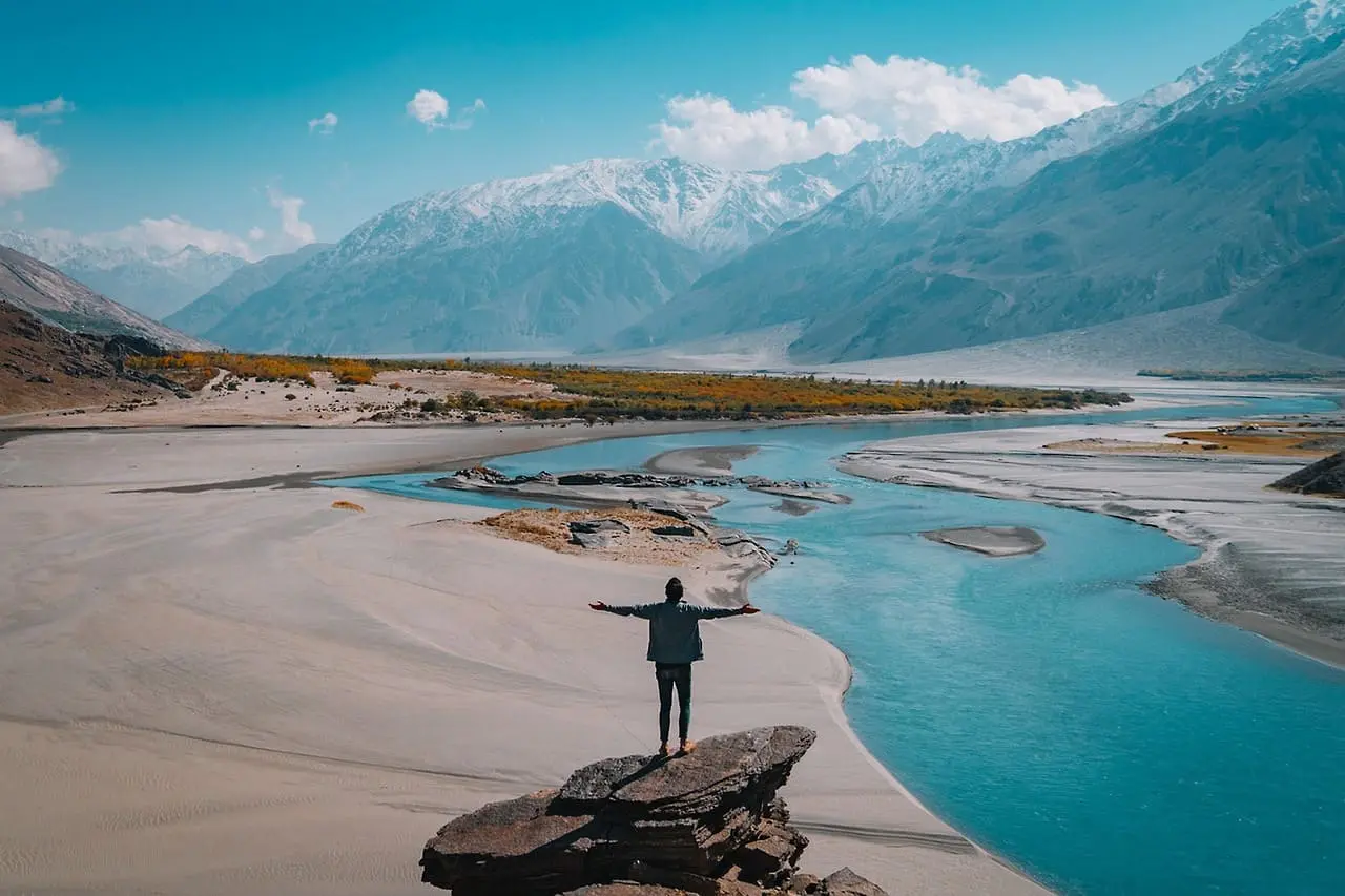 A person standing on a rock with arms outstretched, overlooking a winding blue river surrounded by sandy terrain and towering snow-capped mountains under a clear sky