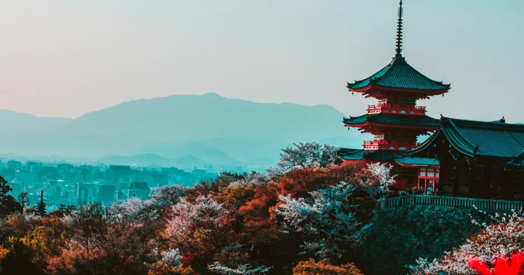 Red and black temple in Japan surrounded by cherry blossoms and colorful trees, with mountains and city skyline in the background. One of the best answers to "what are some good reasons to travel to Japan."